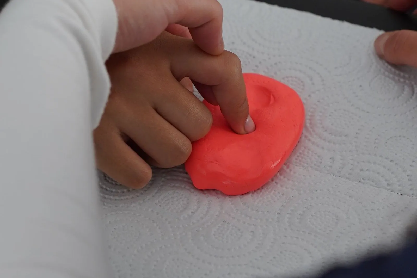 Un copil apasă cu degetul în plastilină roșie, ajutat de mâna unui adult.