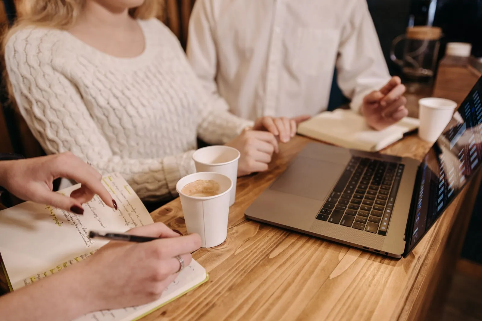 Trei persoane lucrează la o masă de lemn, cu cafea, caiete și un laptop.